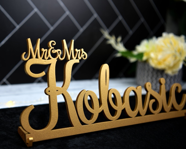 May include: Gold-colored wooden sign with the text "Mr. & Mrs. Kobasic" in a cursive font.