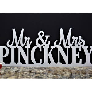 May include: White wooden sign with the text "Mr. & Mrs. PINCKNEY" in a decorative font. The sign is on a dark background with red and pink flowers on either side.