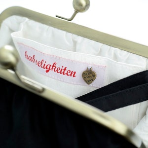 May include: A black fabric coin purse with a silver clasp. The purse is open and the inside is lined with white fabric. A small tag with the text "Made with Love" is attached to the lining. The lining also has a red label with the text "habsseligkeiten".