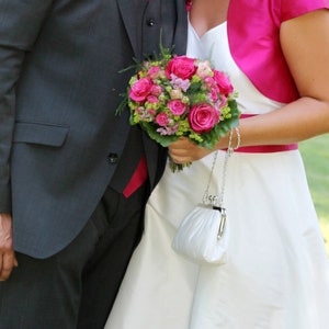 May include: A bride and groom holding a bouquet of pink roses and greenery. The bride is wearing a white dress with a pink bolero jacket and a white clutch.