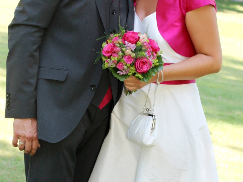 May include: A bride and groom holding a bouquet of pink roses and greenery. The bride is wearing a white dress with a pink bolero jacket and a white clutch.