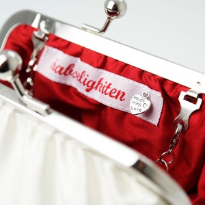 May include: A white satin clutch purse with a silver clasp and chain. The purse is open and the inside is lined with red satin. A small silver tag with the text "Made with Love" is attached to the inside of the purse. The purse has a label that reads "halsigkitten".
