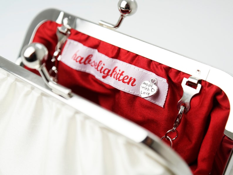 May include: A white satin clutch purse with a silver clasp and chain. The purse is open and the inside is lined with red satin. A small silver tag with the text "Made with Love" is attached to the inside of the purse. The purse has a label that reads "halsigkitten".