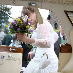 May include: A bride in a white lace dress holds a bouquet of purple, yellow, and white flowers while getting into a vintage car. The car has a wooden interior and a white leather backseat.