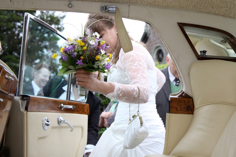 May include: A bride in a white lace dress holds a bouquet of purple, yellow, and white flowers while getting into a vintage car. The car has a wooden interior and a white leather backseat.