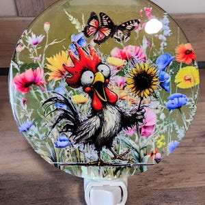 Puede incluir: Una luz nocturna blanca con un inserto de vidrio redondo y colorido que presenta un gallo de dibujos animados sosteniendo un girasol. El gallo tiene una cresta y una papada rojas, plumas blancas y negras y ojos grandes y redondos. El fondo es un campo verde con flores coloridas y una mariposa.