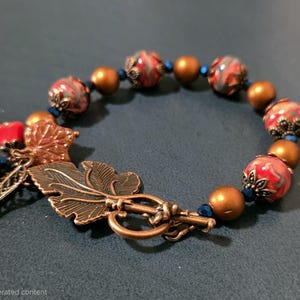 May include: A bracelet featuring round beads in marbled red and gray, solid matte copper-toned spheres, and small blue spacer beads. The piece includes antique copper-toned metal leaf charms and a toggle clasp with a leaf-shaped bar.