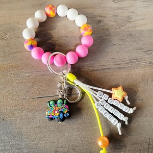 May include: A beaded keychain bracelet with white, pink, and patterned beads. The keychain features a charm of two aliens in a colorful car and a charm that says "GROOVY ALIEN VIBES".