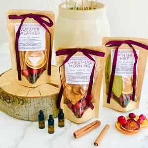 May include: Three kraft paper pouches of simmer pot potpourri labeled Sweater Weather, Christmas Morning, and Winter Hike, each tied with a dark purple ribbon. Includes three small amber glass dropper bottles and two cinnamon sticks on the surface.