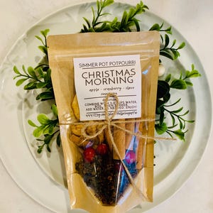 May include: A clear pouch of Christmas Morning simmer pot potpourri featuring dried apple slices, red cranberries, whole cloves, and cinnamon sticks. The label includes directions to combine with 2-3 cups of water and simmer.