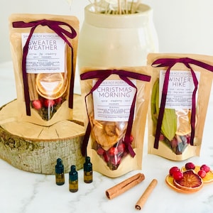 May include: Three kraft paper pouches of simmer pot potpourri labeled Sweater Weather, Christmas Morning, and Winter Hike, each tied with a dark purple ribbon. Includes three small amber glass dropper bottles and two cinnamon sticks on the surface.