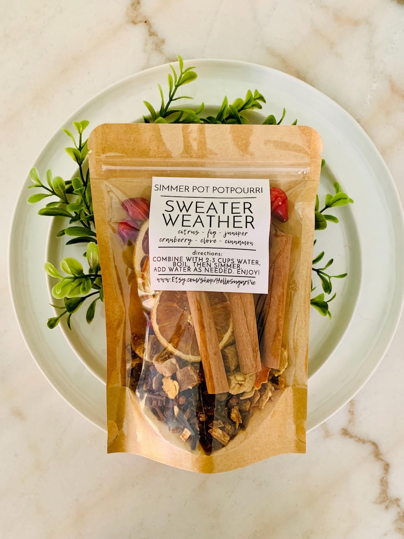 May include: A clear and brown pouch of Sweater Weather simmer pot potpourri containing dried orange slices, cinnamon sticks, and spices. The label lists ingredients including citrus, fig, juniper, cranberry, clove, and cinnamon with instructions.