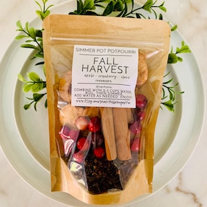 May include: A clear and brown pouch of Fall Harvest simmer pot potpourri containing dried apple slices, red cranberries, cinnamon sticks, and cloves. The label reads: SIMMER POT POTPOURRI, FALL HARVEST, apple, cranberry, clove, cinnamon, directions.