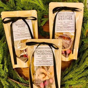 May include: Three beige pouches of simmer pot potpourri tied with black ribbons. Labels read Winter Hike, Sweater Weather, and Christmas Morning, listing ingredients like citrus, fig, cranberry, clove, and cinnamon with boiling instructions.