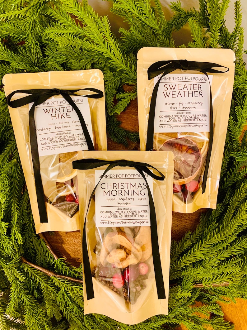 May include: Three beige pouches of simmer pot potpourri tied with black ribbons. Labels read Winter Hike, Sweater Weather, and Christmas Morning, listing ingredients like citrus, fig, cranberry, clove, and cinnamon with boiling instructions.