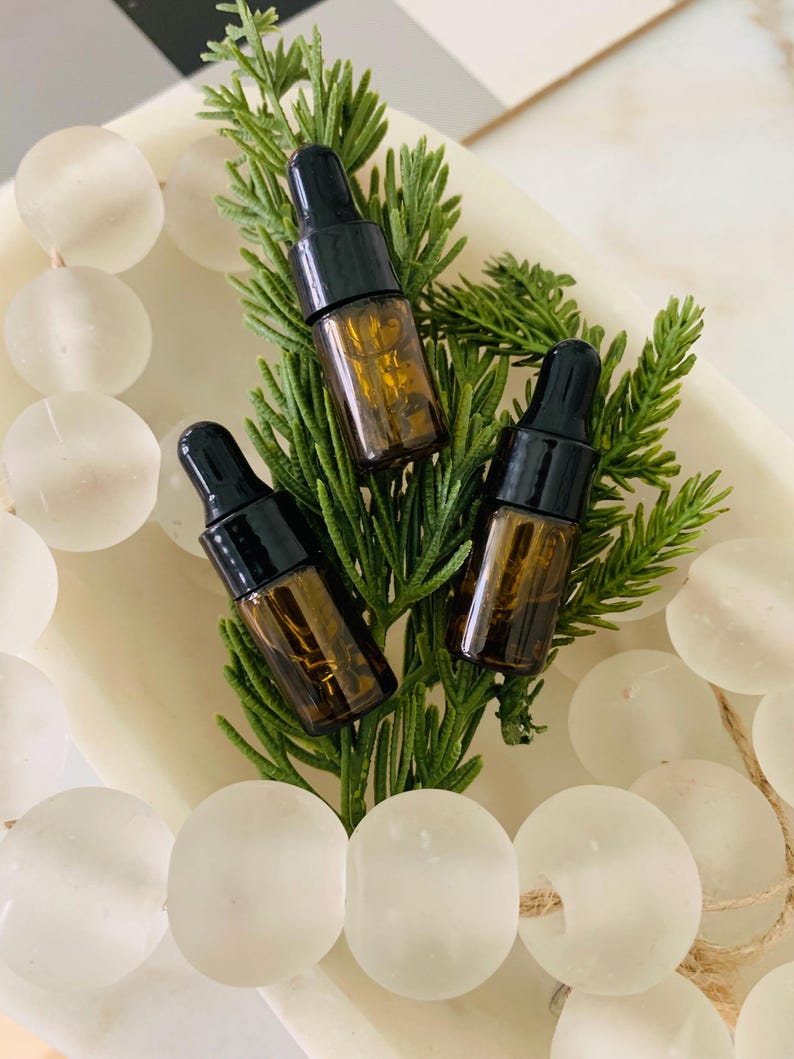 May include: Three small amber glass dropper bottles with black caps are arranged on a green coniferous sprig. The bottles are surrounded by a string of translucent, frosted white glass beads resting in a shallow, off-white oval tray.