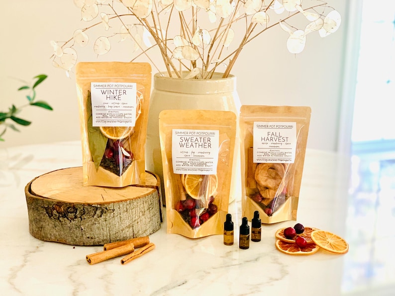 May include: Three stand-up pouches of simmer pot potpourri labeled Winter Hike, Sweater Weather, and Fall Harvest. Each contains dried fruit, spices, and botanicals. Three small amber glass dropper bottles, cinnamon sticks, and dried orange slices are included.