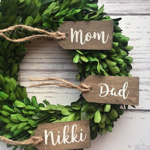 May include: A green boxwood wreath with three wooden tags attached. The tags are tied with twine and have the words "Mom", "Dad", and "Nikki" written in white.