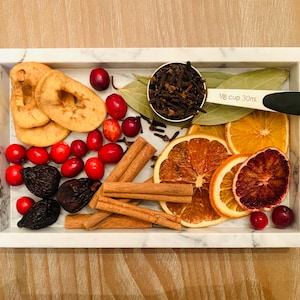 May include: A rectangular white marble tray containing dried apple slices, red cranberries, dark figs, cinnamon sticks, dried orange slices, and bay leaves. A measuring scoop labeled 1/8 cup 30ml holds whole cloves.