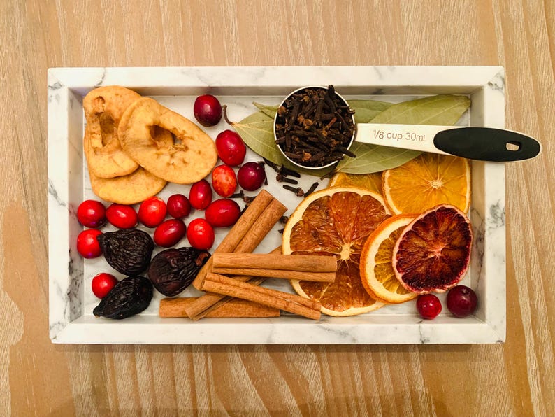 May include: A rectangular white marble tray containing dried apple slices, red cranberries, dark figs, cinnamon sticks, dried orange slices, and bay leaves. A measuring scoop labeled 1/8 cup 30ml holds whole cloves.