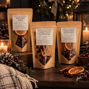 May include: Three brown paper pouches of simmer pot potpourri labeled Winter Hike, Christmas Morning, and Sweater Weather. Each pouch features a clear window displaying dried orange slices, cranberries, and spices. The labels include instructions to combine with 2-3 cups of water and simmer.