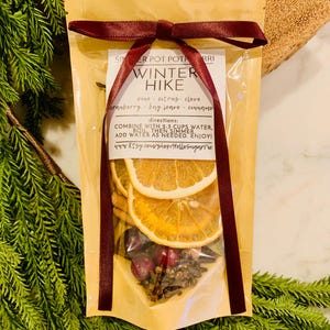 May include: A tan pouch of Winter Hike simmer pot potpourri tied with a dark red ribbon. The label lists ingredients including pine, citrus, clove, cranberry, bay leaves, and cinnamon, with instructions to combine with 2-3 cups of water.