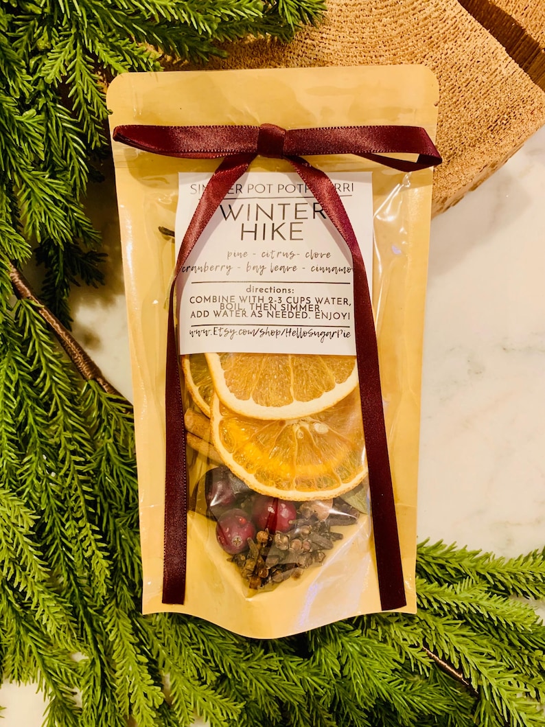 May include: A tan pouch of Winter Hike simmer pot potpourri tied with a dark red ribbon. The label lists ingredients including pine, citrus, clove, cranberry, bay leaves, and cinnamon, with instructions to combine with 2-3 cups of water.
