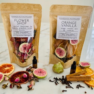 May include: Two stand-up pouches of simmer pot potpourri labeled Flower Shop and Orange Vanilla. The pouches contain dried fruit slices, flower buds, and spices. Small glass dropper bottles and loose dried ingredients are arranged in front.