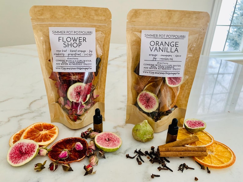 May include: Two stand-up pouches of simmer pot potpourri labeled Flower Shop and Orange Vanilla. The pouches contain dried fruit slices, flower buds, and spices. Small glass dropper bottles and loose dried ingredients are arranged in front.