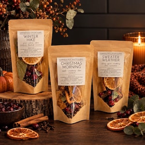 May include: Three brown kraft paper pouches of simmer pot potpourri labeled Winter Hike, Christmas Morning, and Sweater Weather. Each pouch features a clear window revealing dried orange slices, cranberries, and spices inside the packaging.