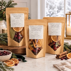 May include: Three brown kraft paper pouches of simmer pot potpourri labeled Winter Hike, Christmas Morning, and Sweater Weather. Each pouch features a clear window displaying dried orange slices, cranberries, and spices with printed instructions.