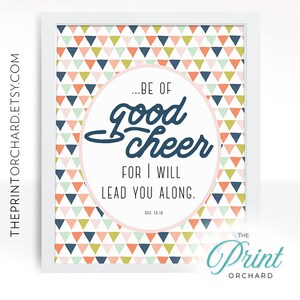 May include: A printable wall art design with a pink and white background featuring a geometric pattern of triangles in shades of blue, green, pink, and orange. The text reads "...BE OF good cheer FOR I WILL LEAD YOU ALONG. DEC 78:18" in a navy blue font. The text is surrounded by a white circle. The bottom right corner of the design features the text "THE Print ORCHARD" in a teal blue font.