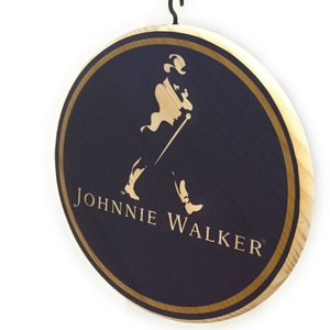 May include: A round wooden sign with a gold border and a black background. The sign features the Johnnie Walker logo, which includes a man in a top hat walking with a cane. The text "JOHNNIE WALKER" is written in gold letters below the logo.
