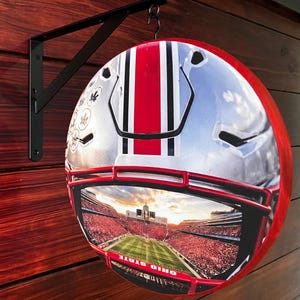 May include: A round sign featuring a football helmet design with a red stripe and the words "OHIO STATE". The sign is mounted on a black metal bracket and hangs against a wooden wall.