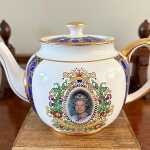 Könnte beinhalten: Weiße Keramik-Teekanne mit goldenen Verzierungen, blauen Akzenten und einem Porträt einer Frau mit Krone. Die Teekanne hat einen goldenen Griff, Ausguss und Deckel. Sie ist mit floralen und Schriftrollenmustern verziert.