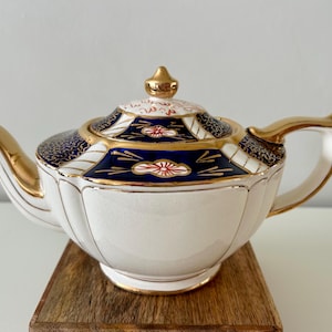 May include: A white teapot with gold trim and blue and red floral designs. The teapot has a gold lid and a gold handle.