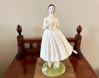May include: A porcelain figurine of a ballerina in a white dress with delicate details. The figurine is standing on a green base with floral accents. The ballerina's pose is graceful, with one leg extended and arms crossed. The figurine is set on a wooden surface.