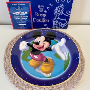 Può includere: Un piatto decorativo della collezione Mickey Mouse, con Topolino su sfondo blu e bordo dorato. Il piatto ha un certificato di autenticità e una scatola Royal Doulton sullo sfondo.