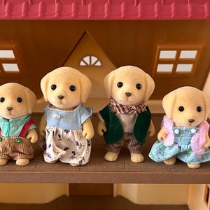 May include: Four toy Labrador Retriever figures, each dressed in miniature outfits, stand on a wooden shelf inside a dollhouse. The figures have tan fur and black eyes. The outfits include overalls, dresses, and vests in various colors.