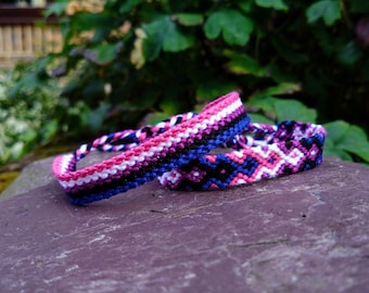 Op de afbeelding: Twee vriendschapsarmbanden gemaakt met kleurrijk borduurgaren. Een armband is roze, wit, blauw en zwart. De andere armband is paars, wit, blauw en zwart.