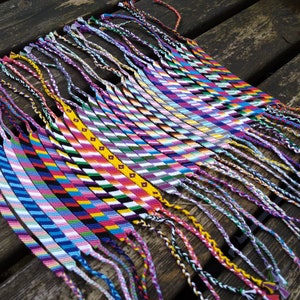 Puede incluir: Una colecci&oacute;n de pulseras de amistad coloridas con varios patrones y colores, incluyendo rosa, azul, verde, amarillo y blanco. Las pulseras est&aacute;n atadas juntas y tienen largas borlas.