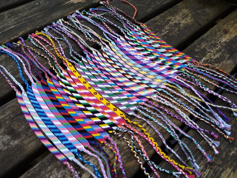 Puede incluir: Una colecci&oacute;n de pulseras de amistad coloridas con varios patrones y colores, incluyendo rosa, azul, verde, amarillo y blanco. Las pulseras est&aacute;n atadas juntas y tienen largas borlas.