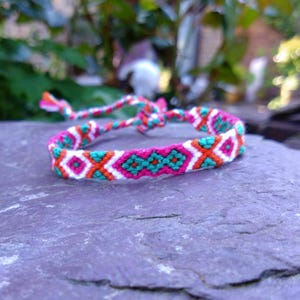 Könnte beinhalten: Ein farbenfrohes Freundschaftsarmband mit einem gewebten Muster. Das Armband zeigt ein sich wiederholendes Design aus weißen, türkisfarbenen, orangefarbenen und rosa Rauten und Kreuzen. Es ist mit einer passenden Kordel gebunden.