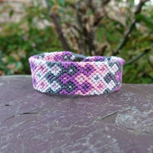 Puede incluir: Un brazalete de amistad con un patrón geométrico en rosa, morado, blanco y gris.