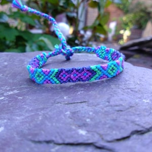 May include: A handmade friendship bracelet in shades of blue, green, and purple. The woven bracelet features a geometric pattern and adjustable ties. The bracelet is displayed on a textured, purple surface.