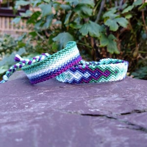 May include: Two friendship bracelets with a green, white and purple colour scheme. The bracelets are woven with a chevron pattern.