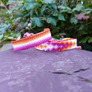 Puede incluir: Dos pulseras de amistad en colores rosa, naranja y blanco. Una pulsera tiene un patrón de chevron y la otra tiene un patrón de rayas simple.