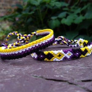May include: Two friendship bracelets in yellow, white, purple, and black colours. One bracelet has a striped pattern, and the other has a diamond pattern.