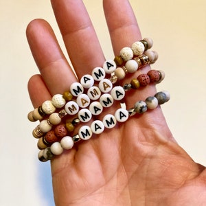 May include: Four bracelets with white beads that spell out "MAMA" in black and gold lettering. The bracelets are made with different types of beads, including lava rock, wood, and white stone.