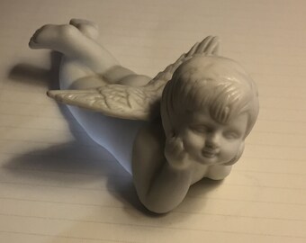 Może przedstawiać: Biała porcelanowa figurka cherubina z skrzydłami leżąca na boku, patrząca w prawo z ręką na brodzie.